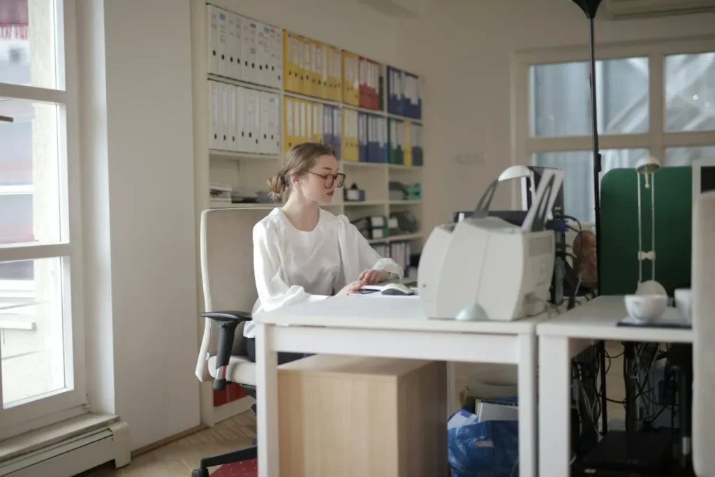 Perempuan bekerja di meja kantor yang penuh berkas dan peralatan, menggambarkan pekerjaan admin general